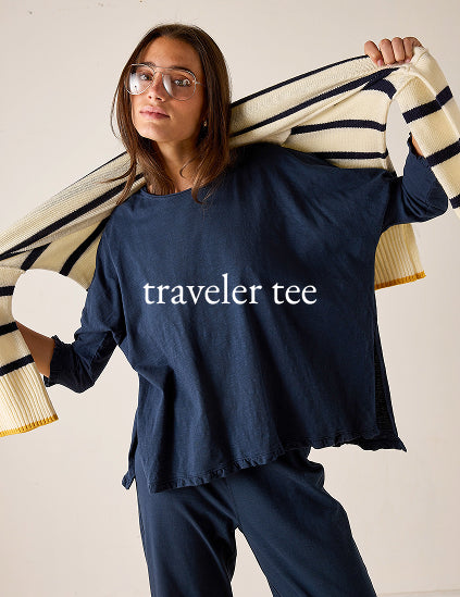 Person wearing a navy 'traveler tee' with a beige and black striped sweater over their shoulder on a light background.