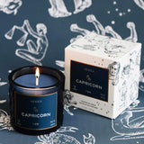 Mersea Capricorn scented candle displayed beside its accompanying box, set against an astrology sign-themed backdrop