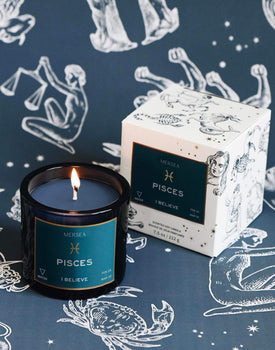 Mersea Pisces scented candle displayed beside its accompanying box, set against an astrology sign-themed backdrop