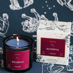 Mersea Scorpio scented candle displayed beside its accompanying box, set against an astrology sign-themed backdrop