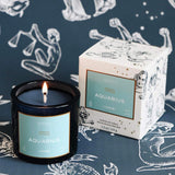 Mersea Aquarius scented candle displayed beside its accompanying box, set against an astrology sign-themed backdrop