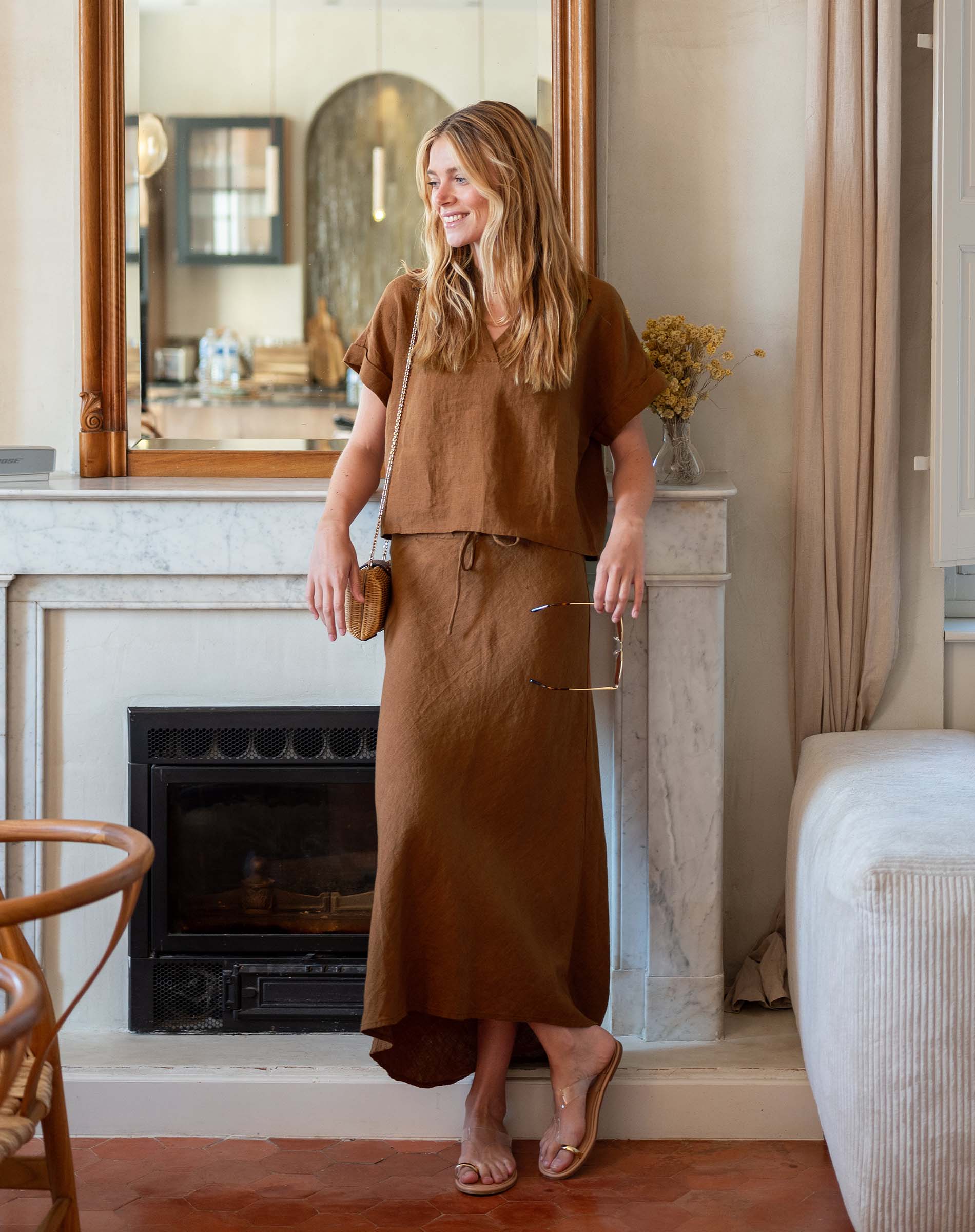 Woman in a brown outfit standing in a room with a fireplace and mirror.