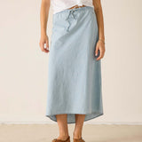 Person wearing a light blue denim skirt with a white top and brown sandals on a plain background