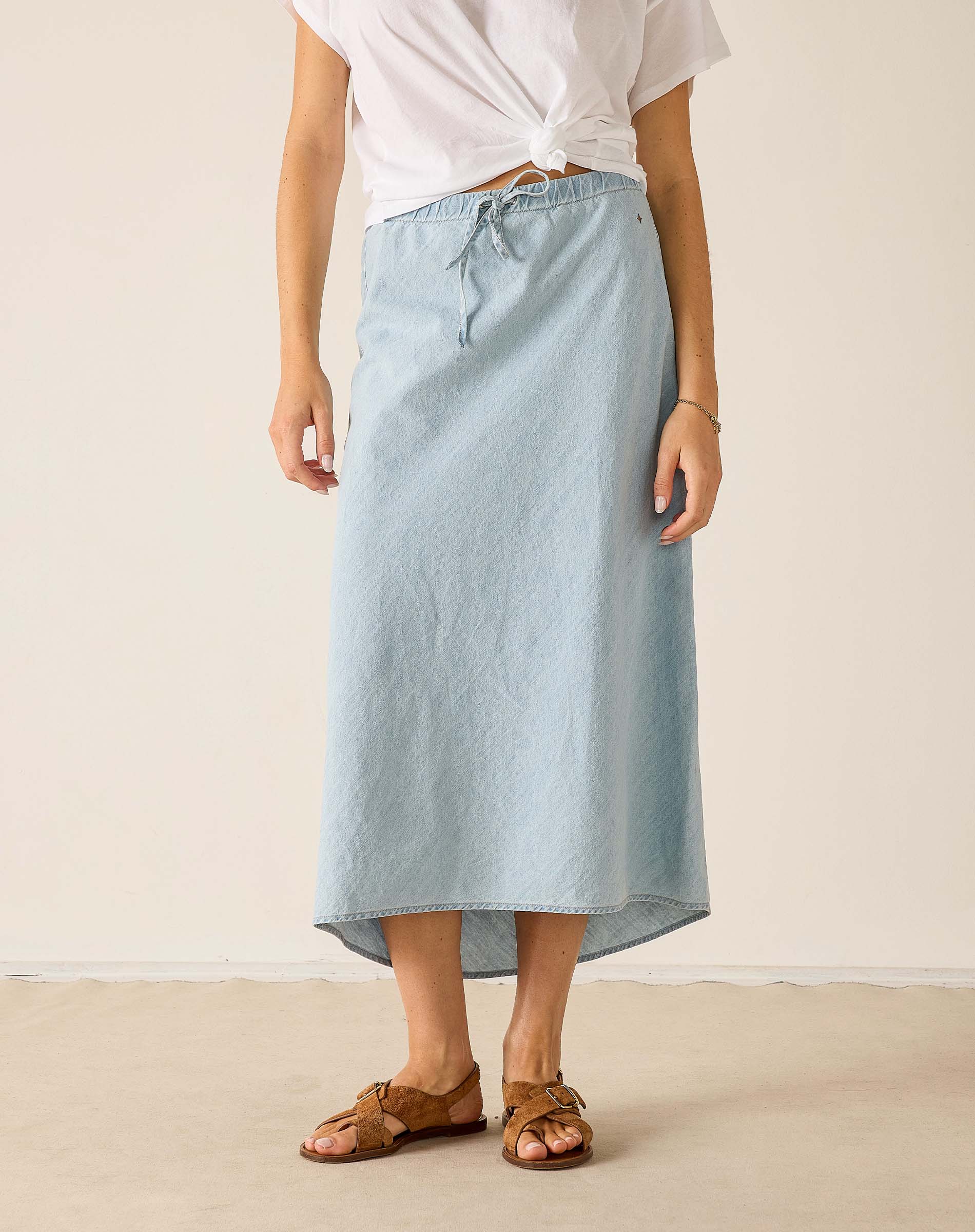 Person wearing a light blue denim skirt with a white top and brown sandals on a plain background