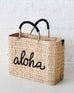 straw medium sized bag with black leather handles and aloha on the front on a white cement ground