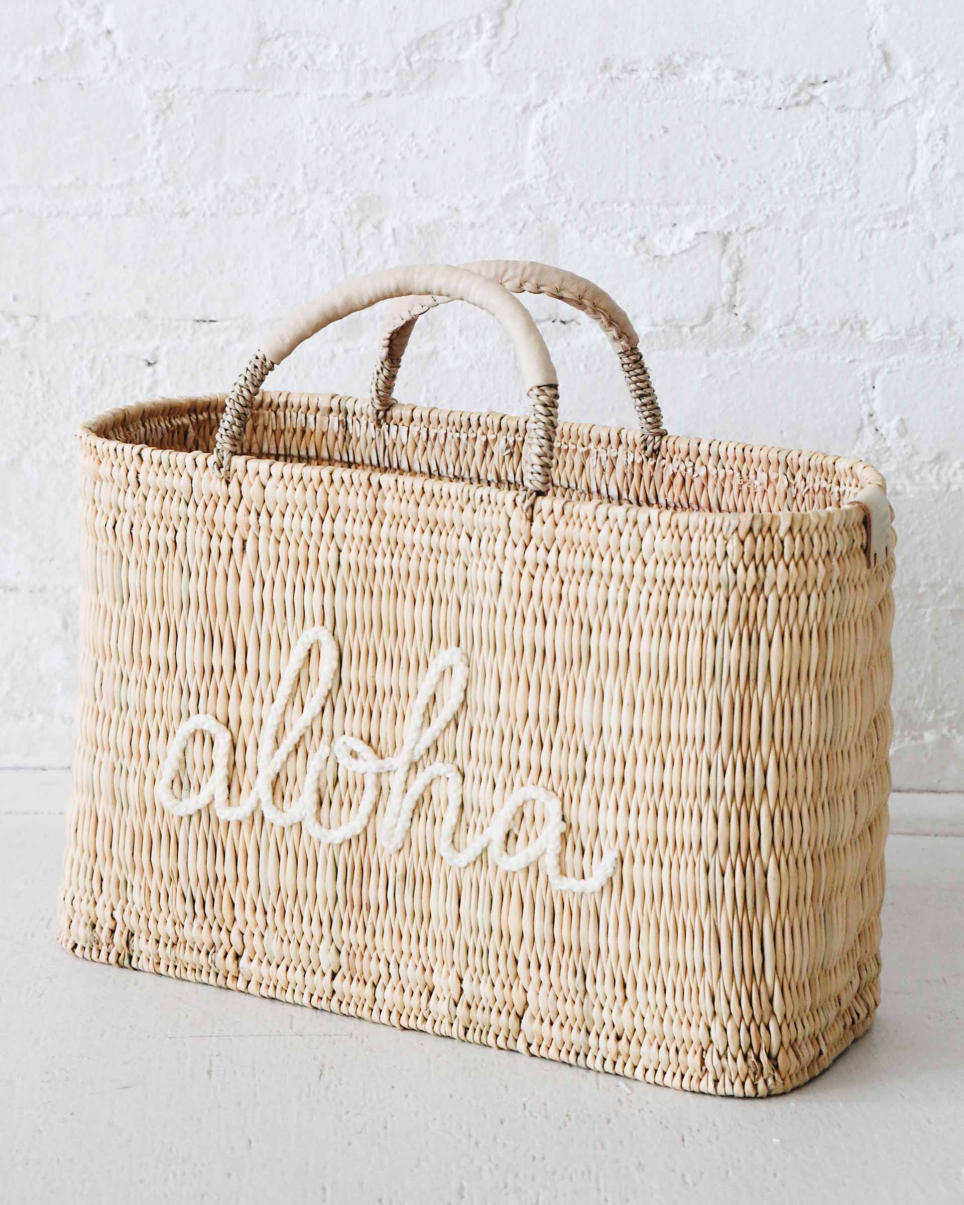 straw medium sized bag with white leather handles and aloha on the front on a white cement ground