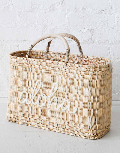 straw medium sized bag with white leather handles and aloha on the front on a white cement ground