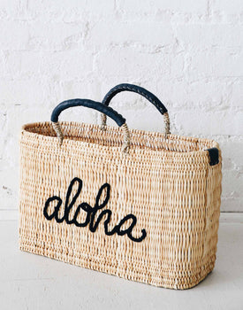 straw medium sized bag with indigo leather handles and aloha on the front on a white cement ground