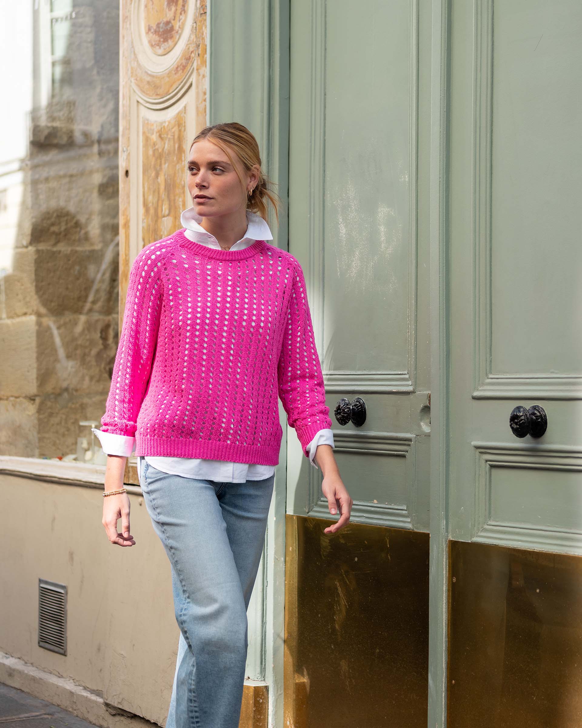 Woman wearing a pink sweater and light blue jeans standing in front of a building entrance.