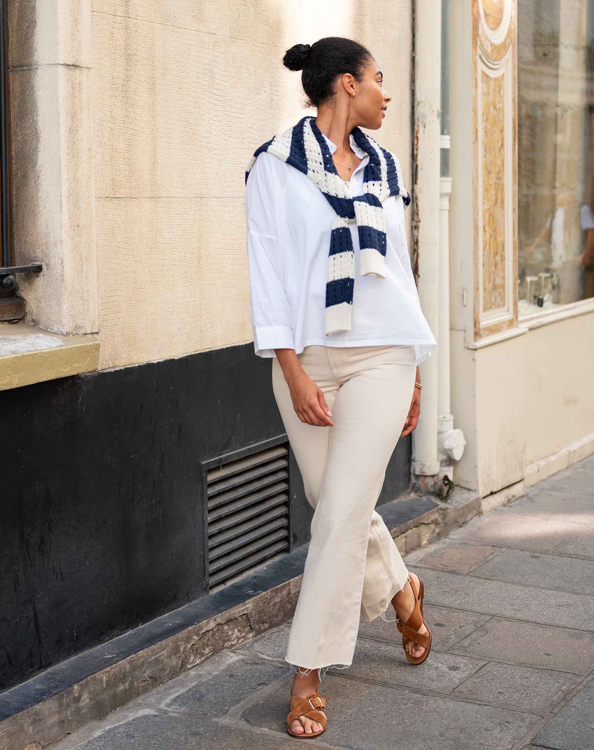Woman wearing a blue and white striped scarf, white shirt, beige pants, and brown sandals on a city street.