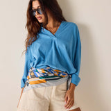 Woman wearing a blue sweater with a colorful pattern over a white shirt, beige shorts, and sunglasses on a plain background.