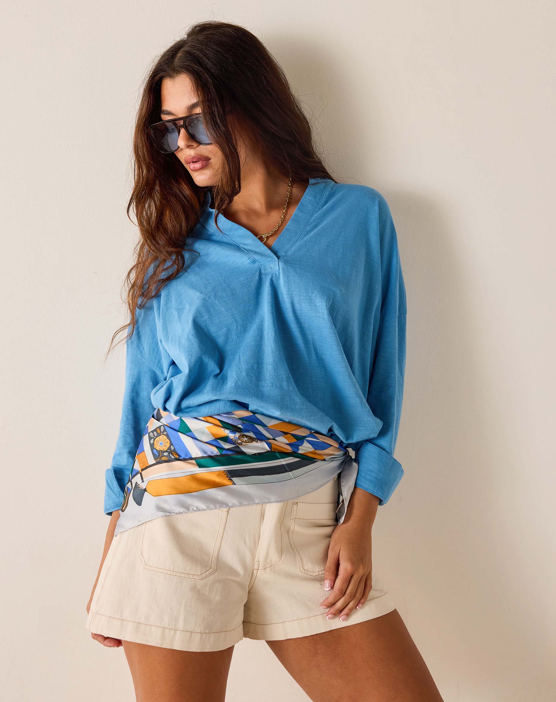 Woman wearing a blue sweater with a colorful pattern over a white shirt, beige shorts, and sunglasses on a plain background.