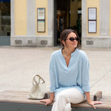 Woman sitting on a bench wearing sunglasses and a light blue top with white pants, with a building in the background.