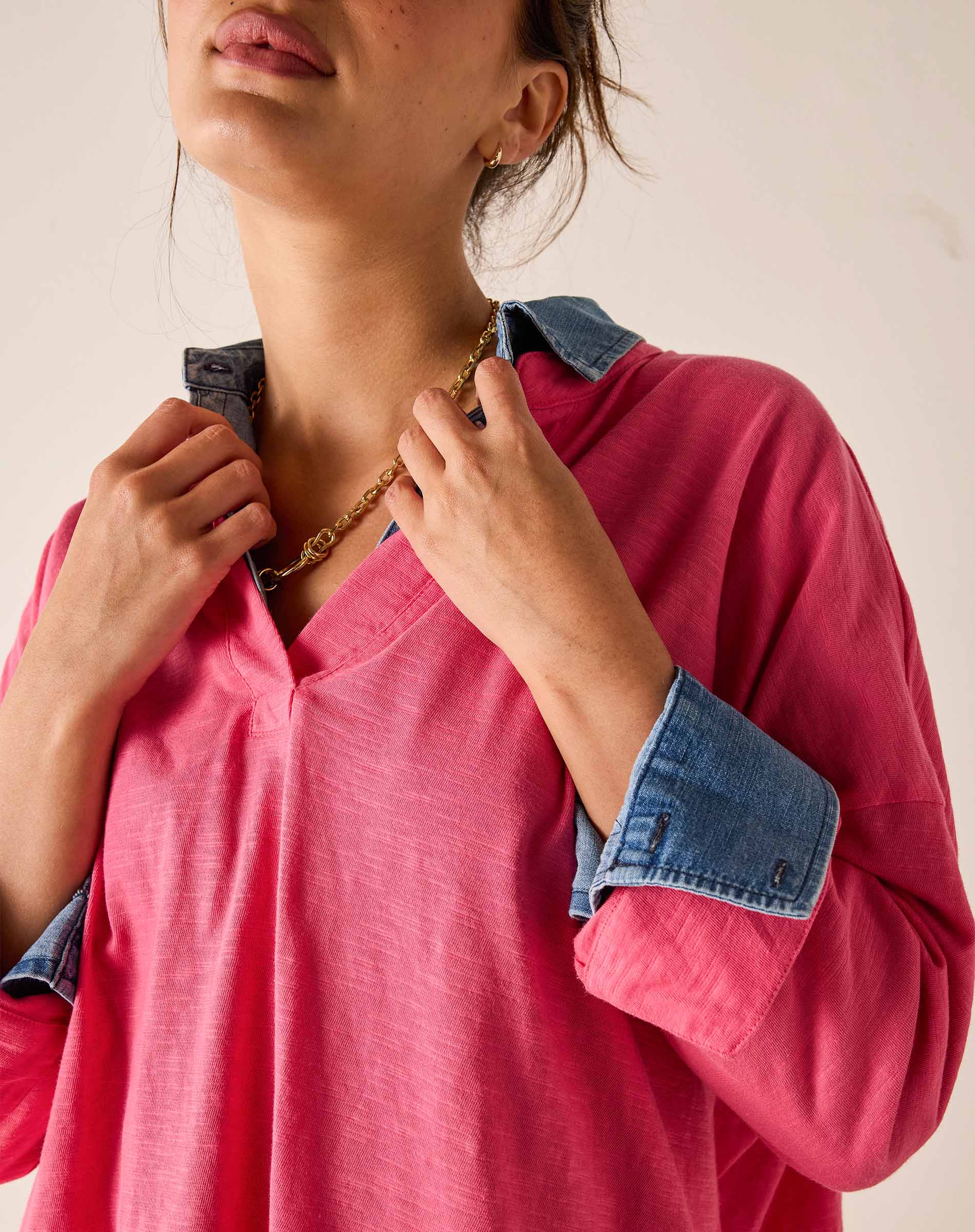 Person wearing a pink shirt with rolled-up denim sleeves, holding a gold chain necklace.