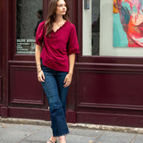 Woman in a red shirt and blue jeans standing on a sidewalk with a colorful painting in the background.