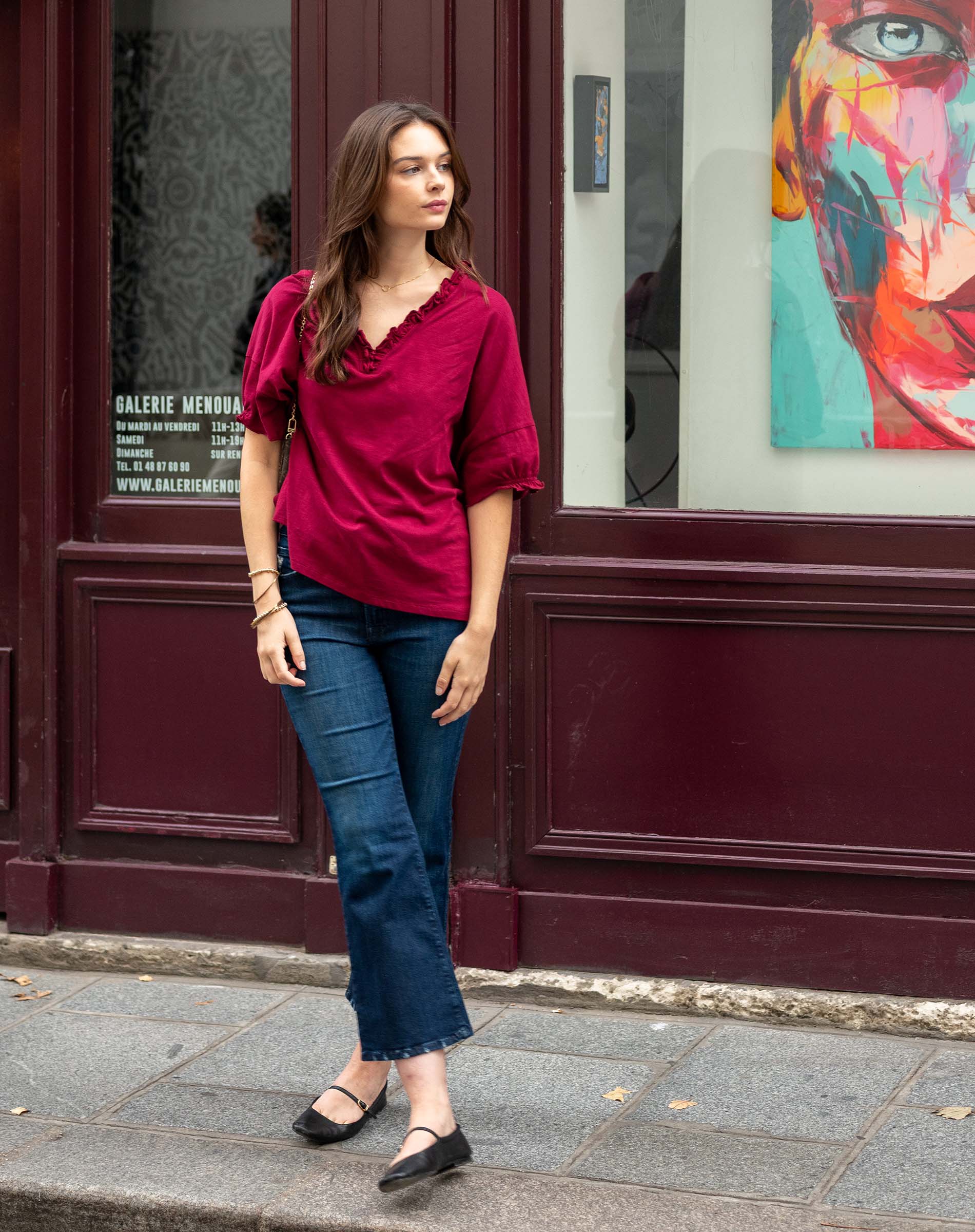 Woman in a red shirt and blue jeans standing on a sidewalk with a colorful painting in the background.