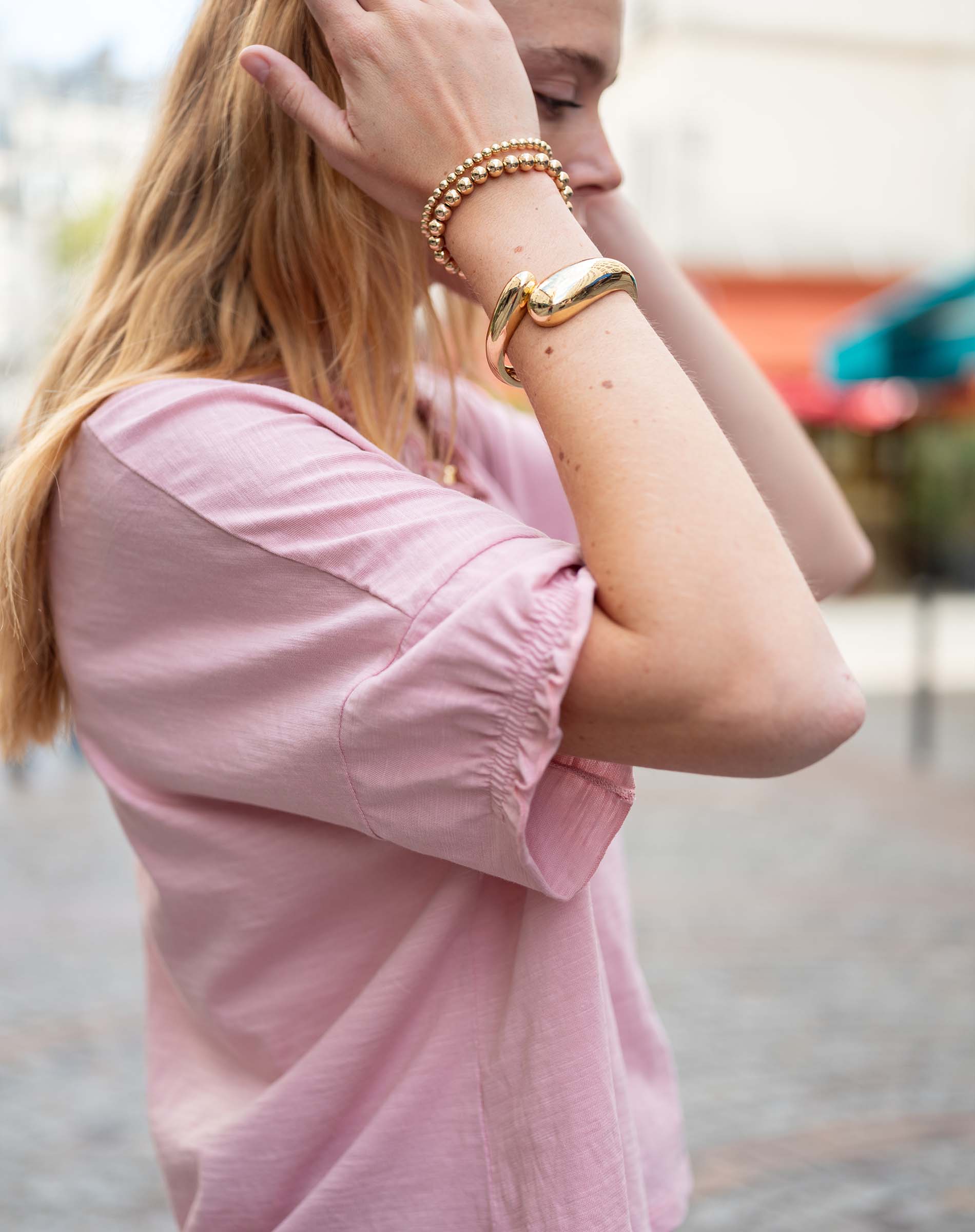 Person wearing a pink shirt with rolled-up sleeves and gold bracelets on a blurred background
