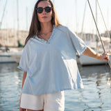 Woman in light blue top and white shorts standing on a boat with sailboats in the background.