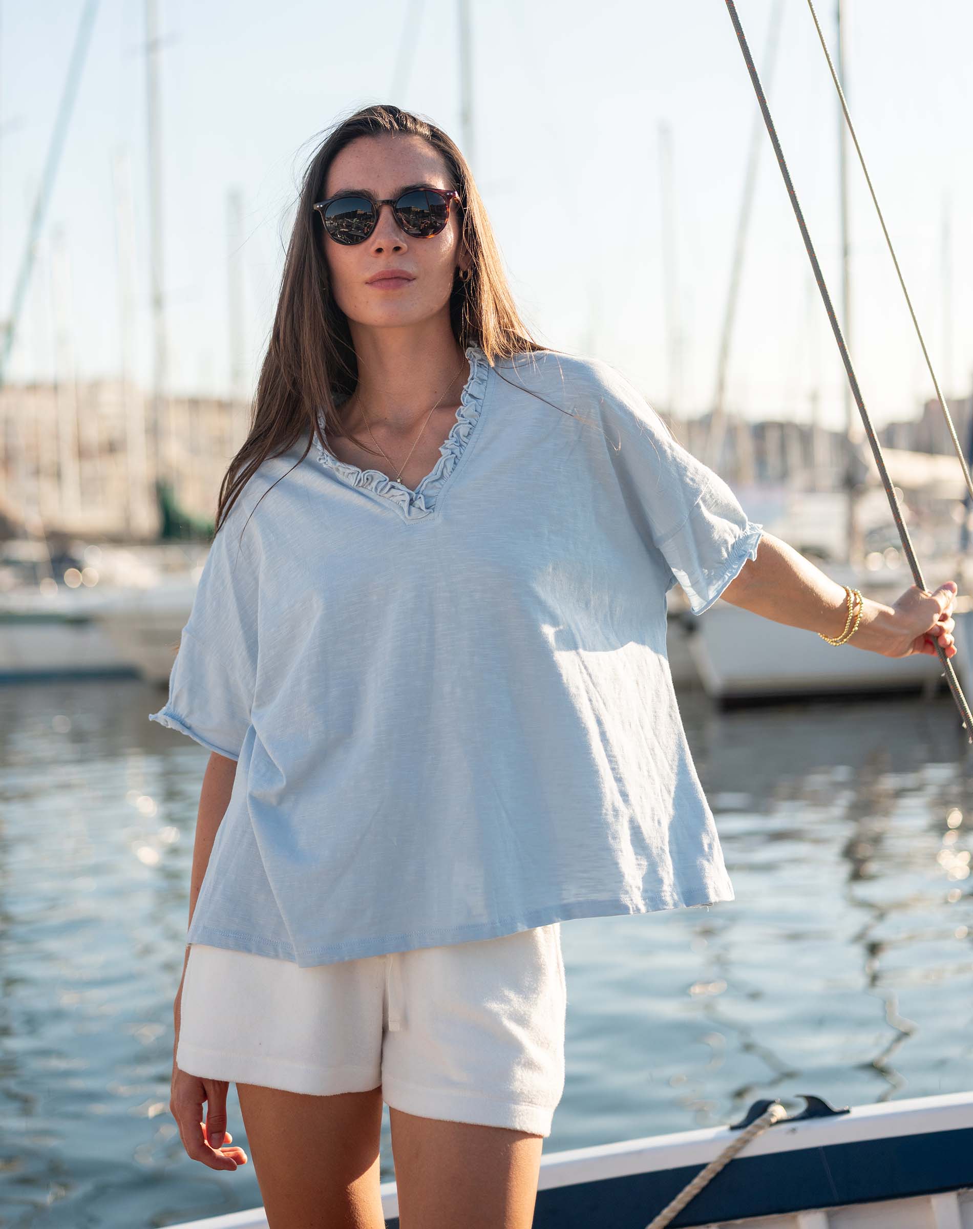 Woman in light blue top and white shorts standing on a boat with sailboats in the background.