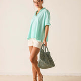Woman wearing a light blue shirt and white shorts, holding a green bag against a plain background