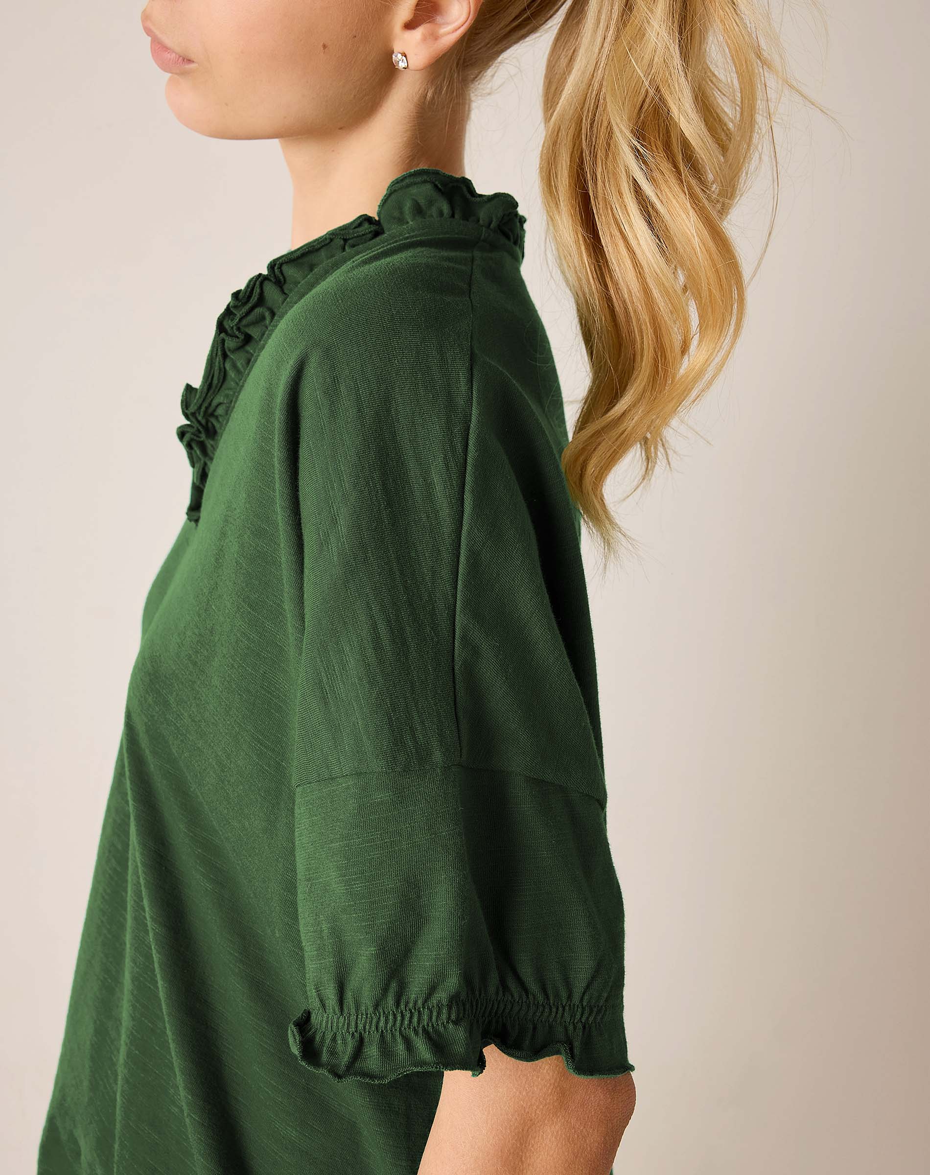 Model in a pine green ruffle tee with ruffled edges along the neckline and sleeve cuffs.