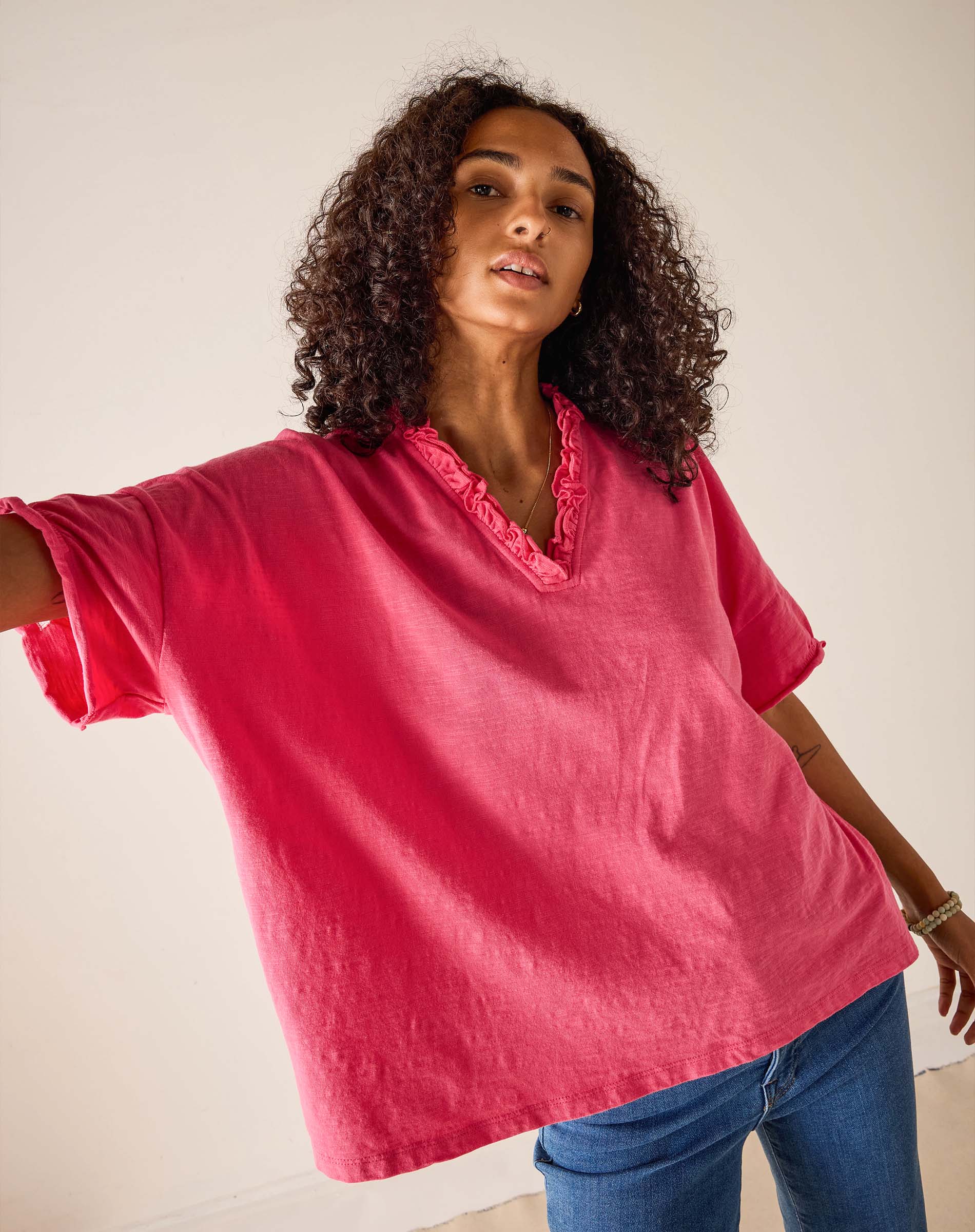 Woman wearing a pink blouse with ruffled neckline and blue jeans against a plain background