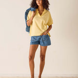 Woman wearing a yellow top and blue shorts holding a denim jacket against a white background