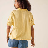 Woman wearing a yellow blouse with ruffled details and denim shorts on a beige background