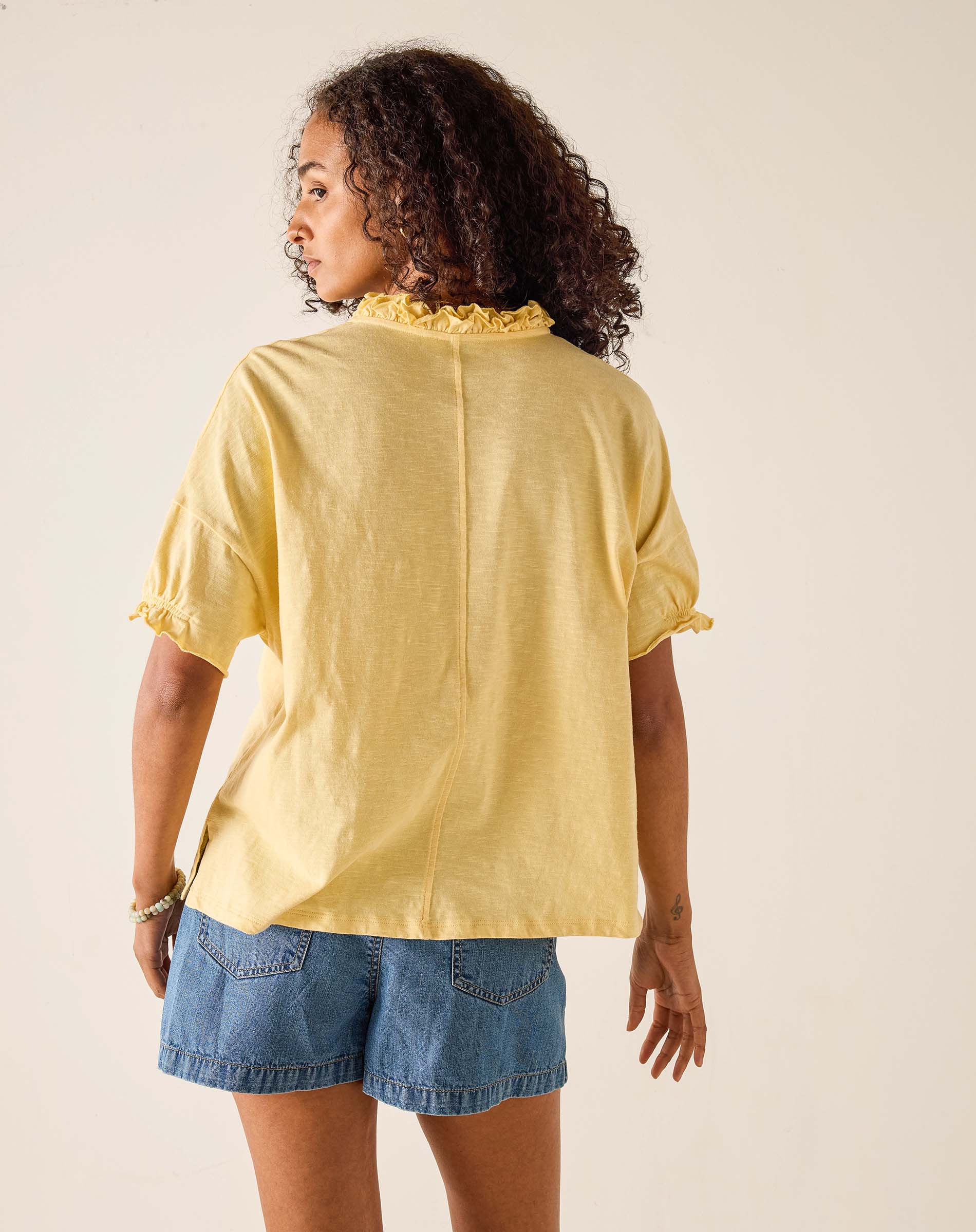 Woman wearing a yellow blouse with ruffled details and denim shorts on a beige background