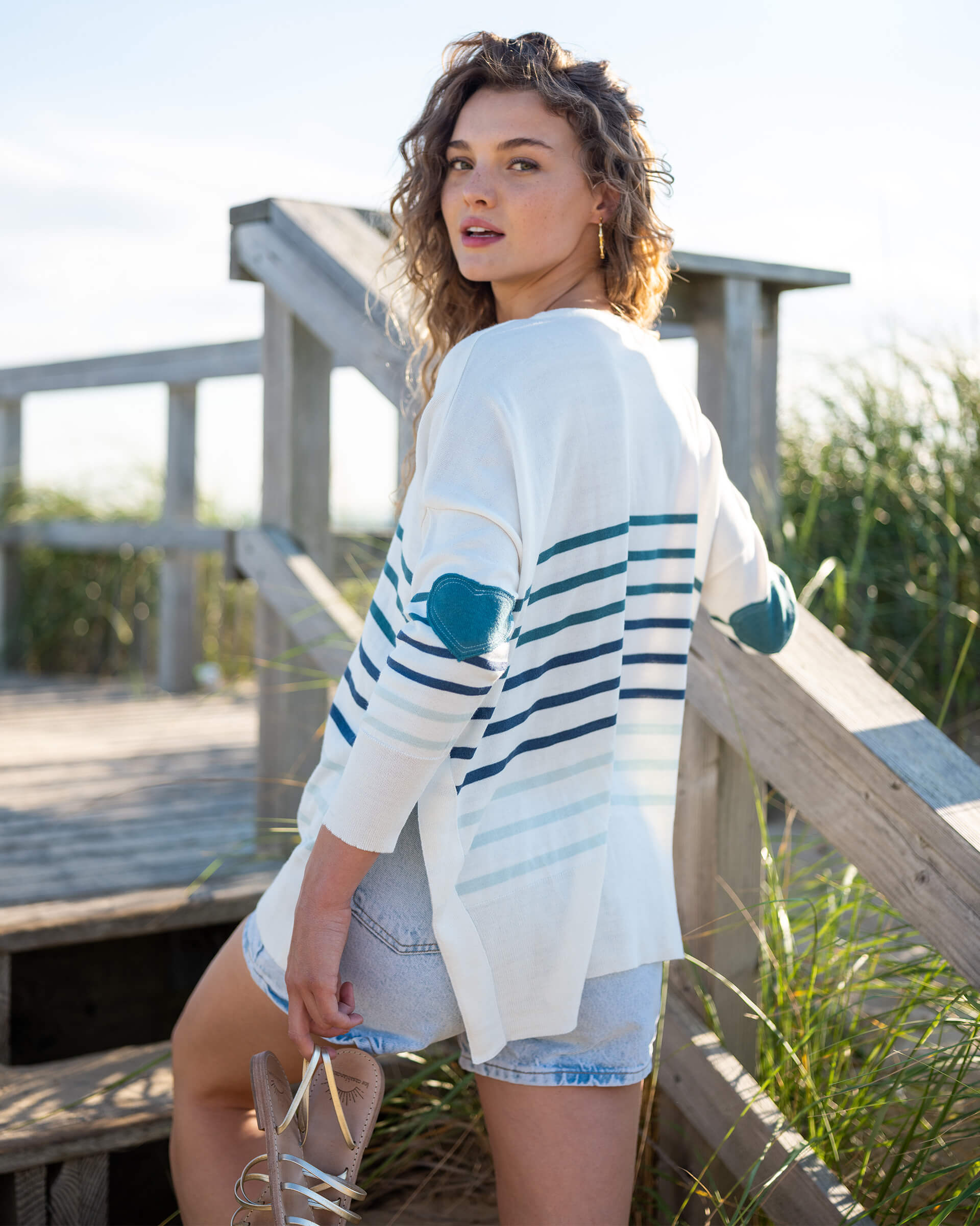female wearing a white sweater with blue stripes and blue heart walking up stairs on the beach