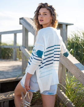 female wearing white sweater with greenish blue stripes and heart elbow patch on a beach