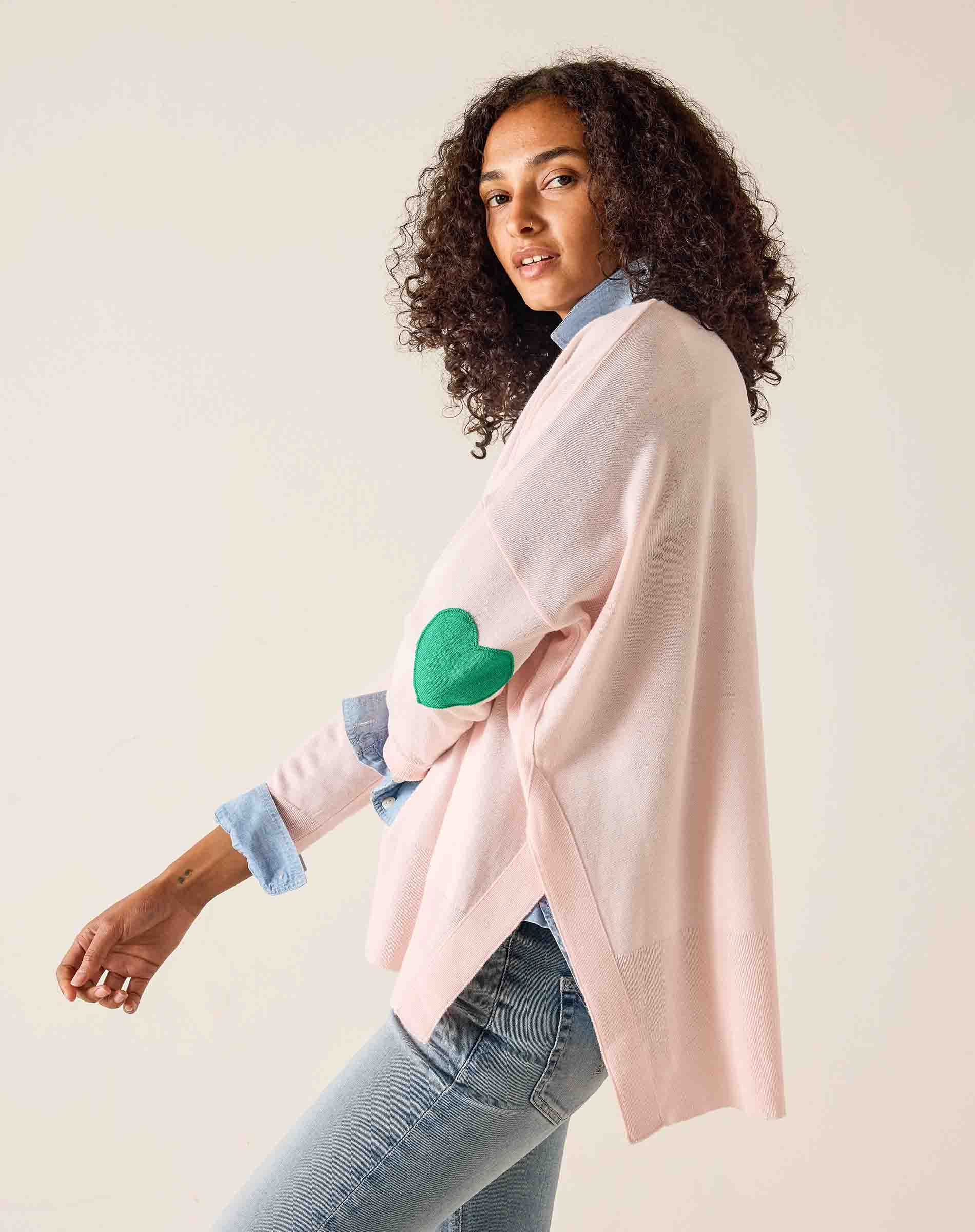 Woman wearing a light pink poncho with green heart elbow patches on a plain background