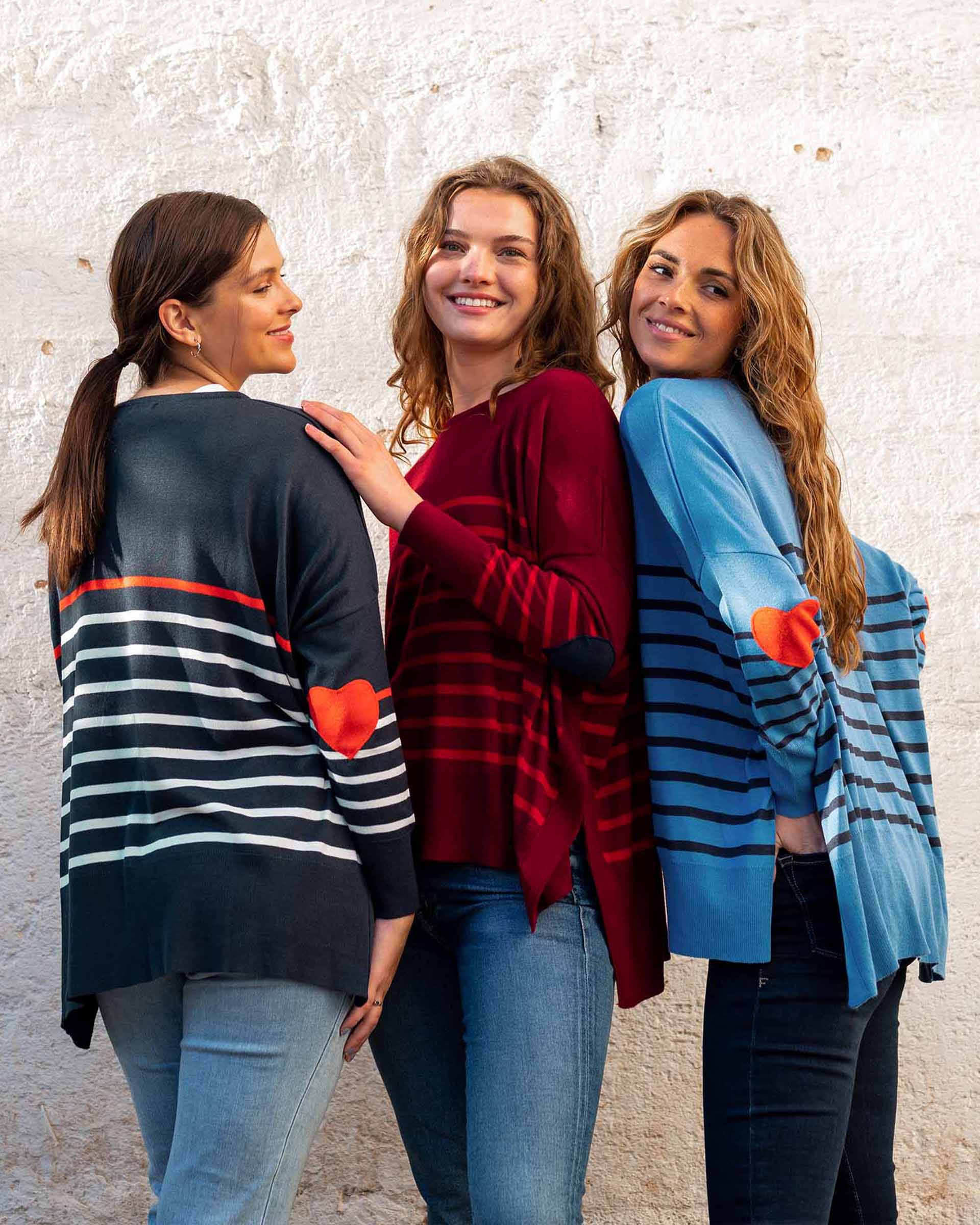 three females wearing navy, red, and blue striped sweaters on a white wall with heart elbow patches