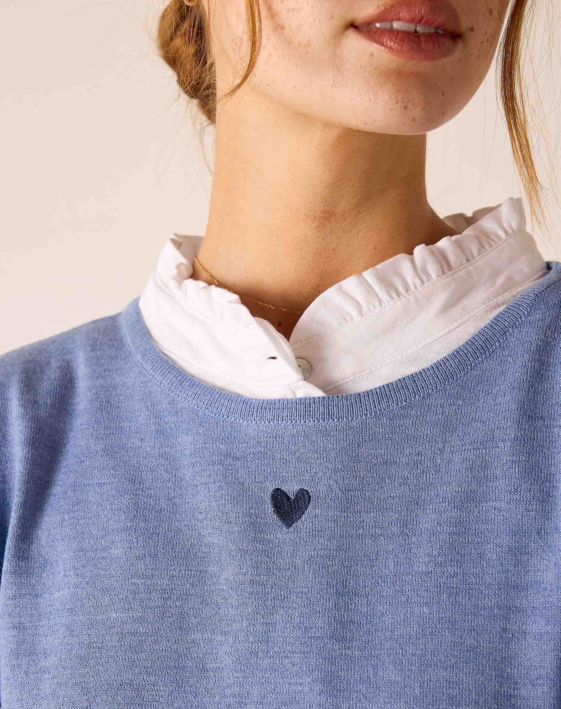 Ocean blue knit sweater with a small heart embroidered on the front and layered over a white ruffled shirt.