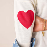 White knit sweater featuring a large red heart graphic on the sleeve.