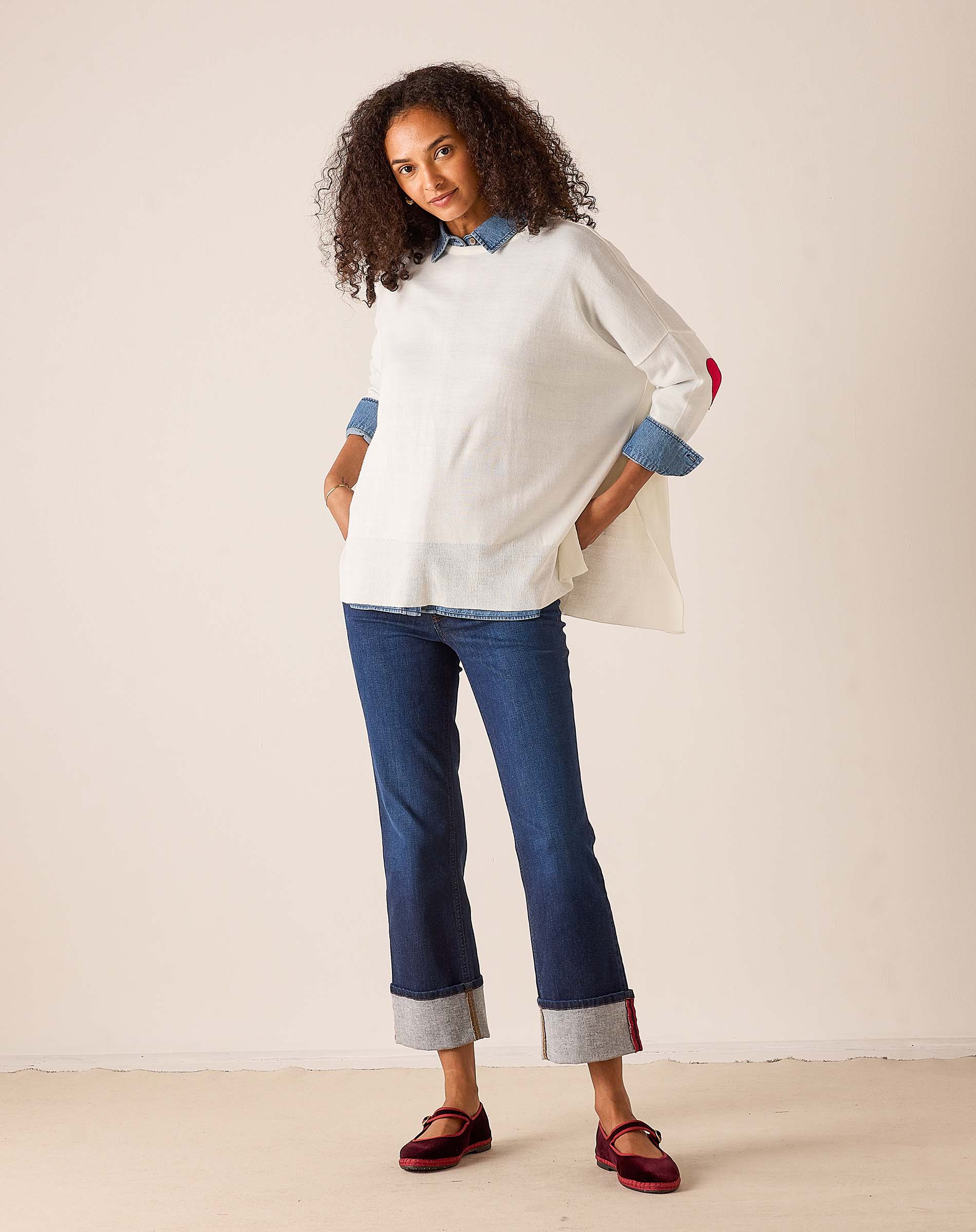 White oversized knit sweater with red accent and layered blue striped shirt, styled with cuffed denim jeans and red loafers.