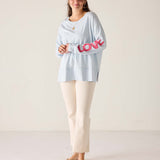 Woman wearing a light blue top with pink text and white pants on a plain background