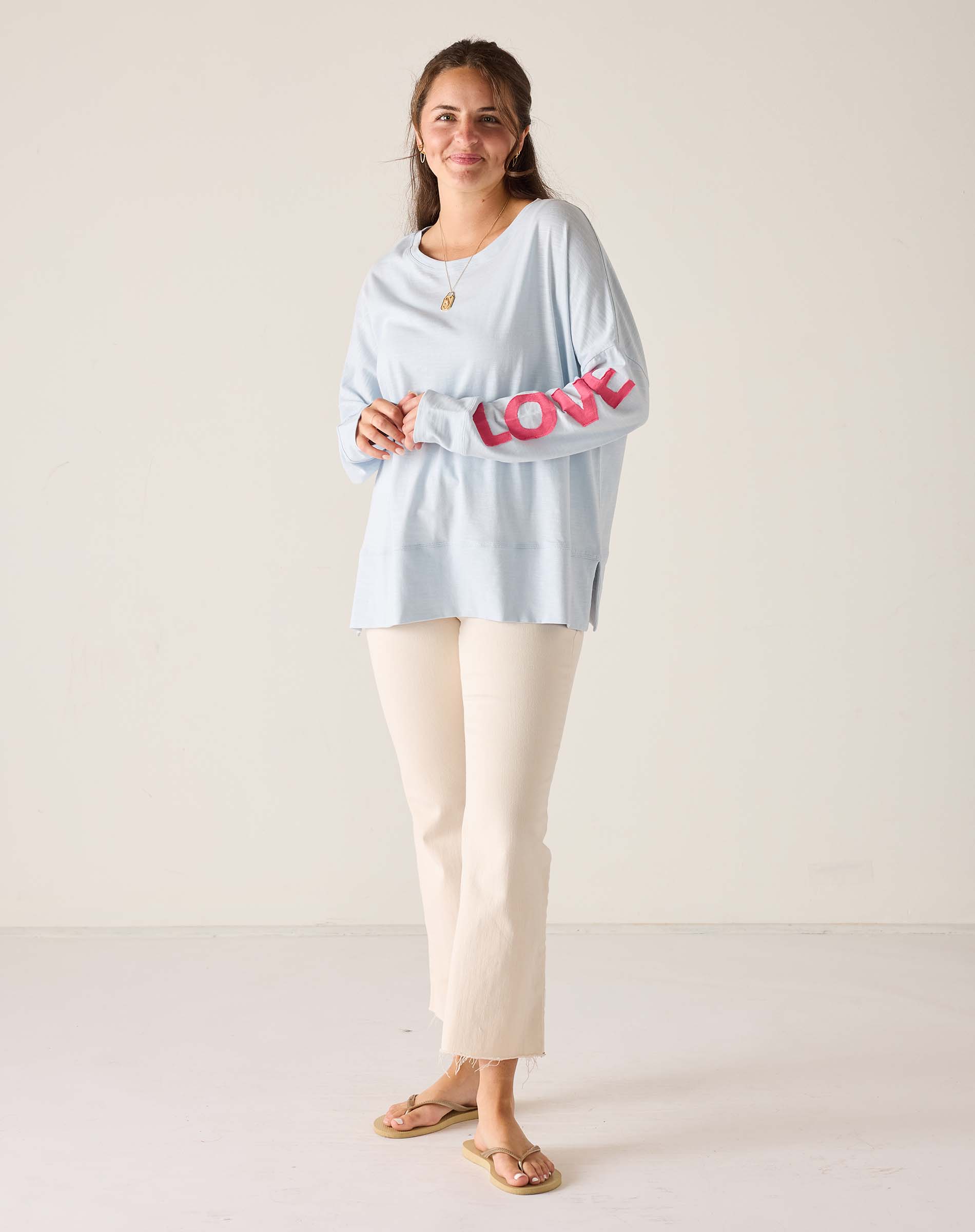 Woman wearing a light blue top with pink text and white pants on a plain background