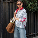 Woman holding a brown bag with flowers, wearing a light gray sweatshirt with red text and blue jeans, standing against a black metal fence.