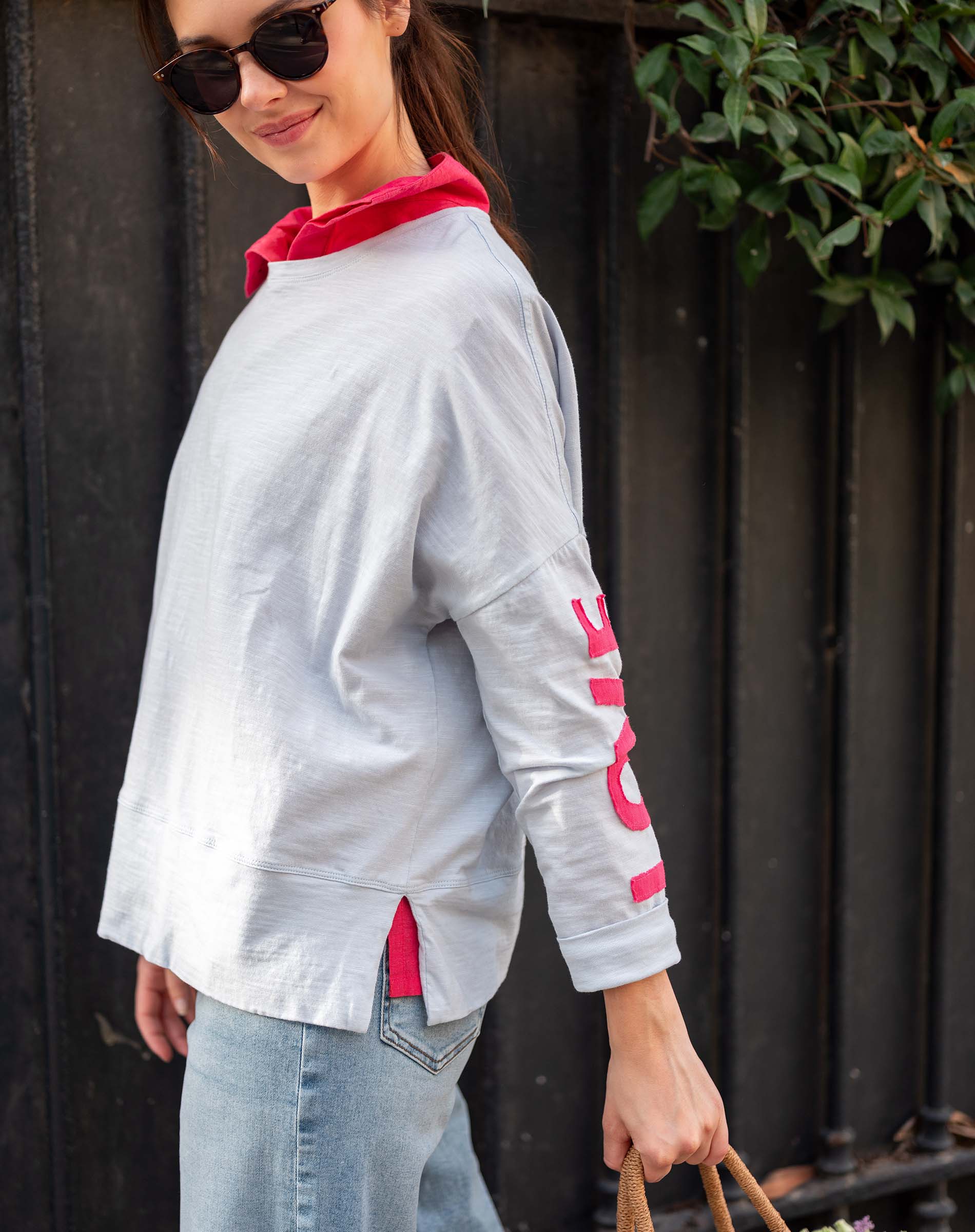 Woman wearing a gray sweatshirt with red accents and sunglasses, standing against a dark background.