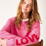 Woman wearing a pink sweatshirt with 'LOVE' on it, sitting on a wooden chair against a white background