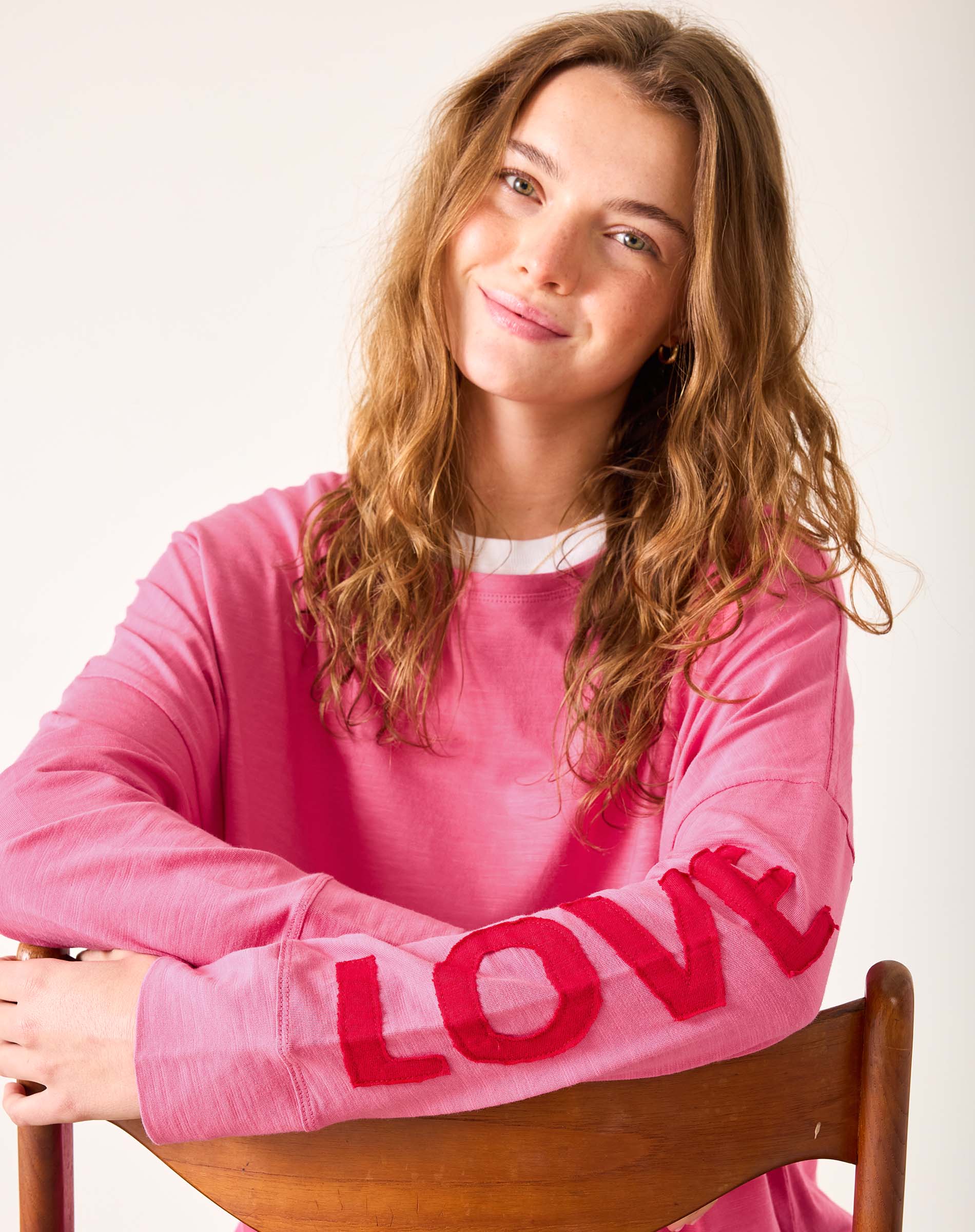 Woman wearing a pink sweatshirt with 'LOVE' on it, sitting on a wooden chair against a white background