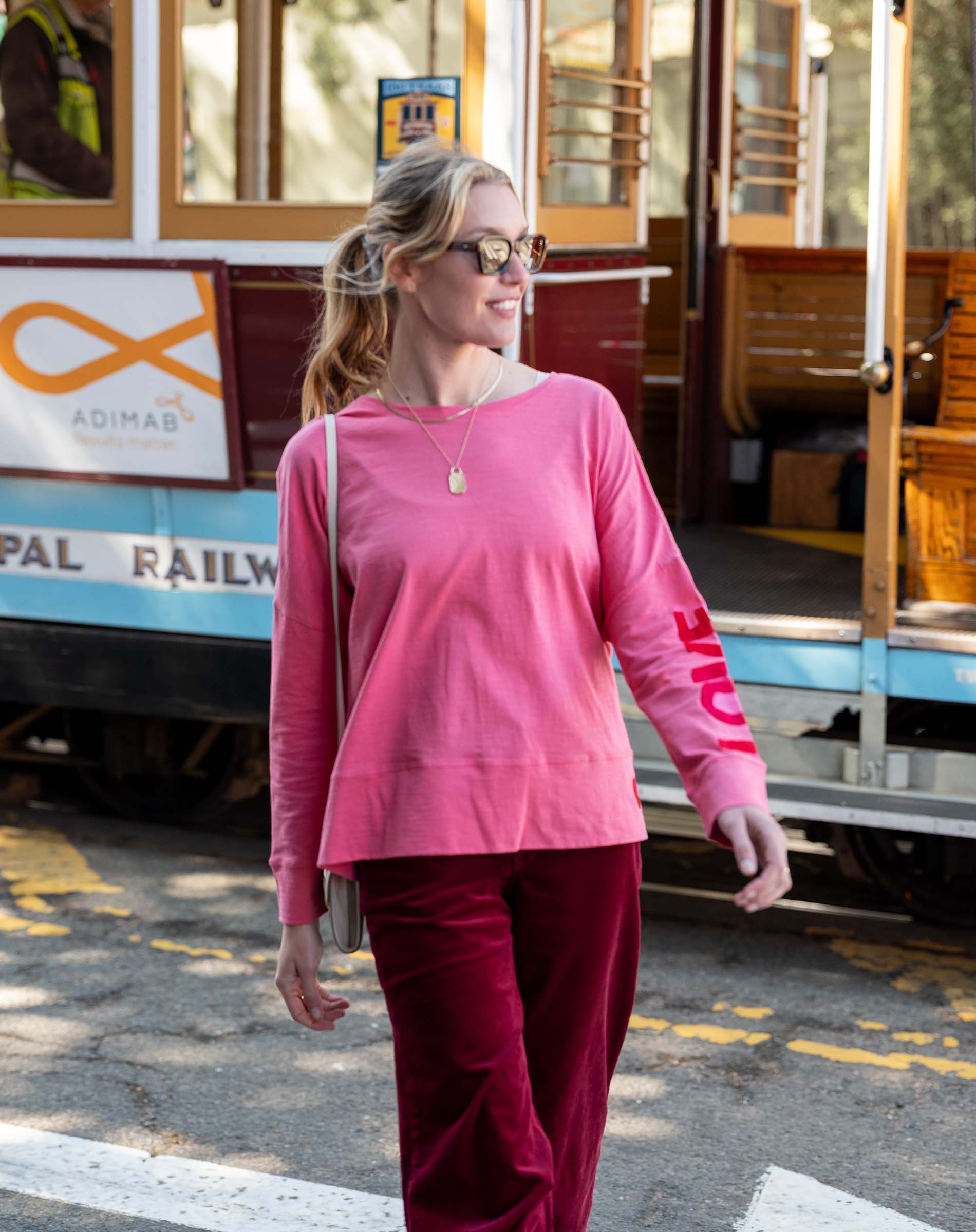 Woman in pink long-sleeve top and red pants walking past a trolley car.