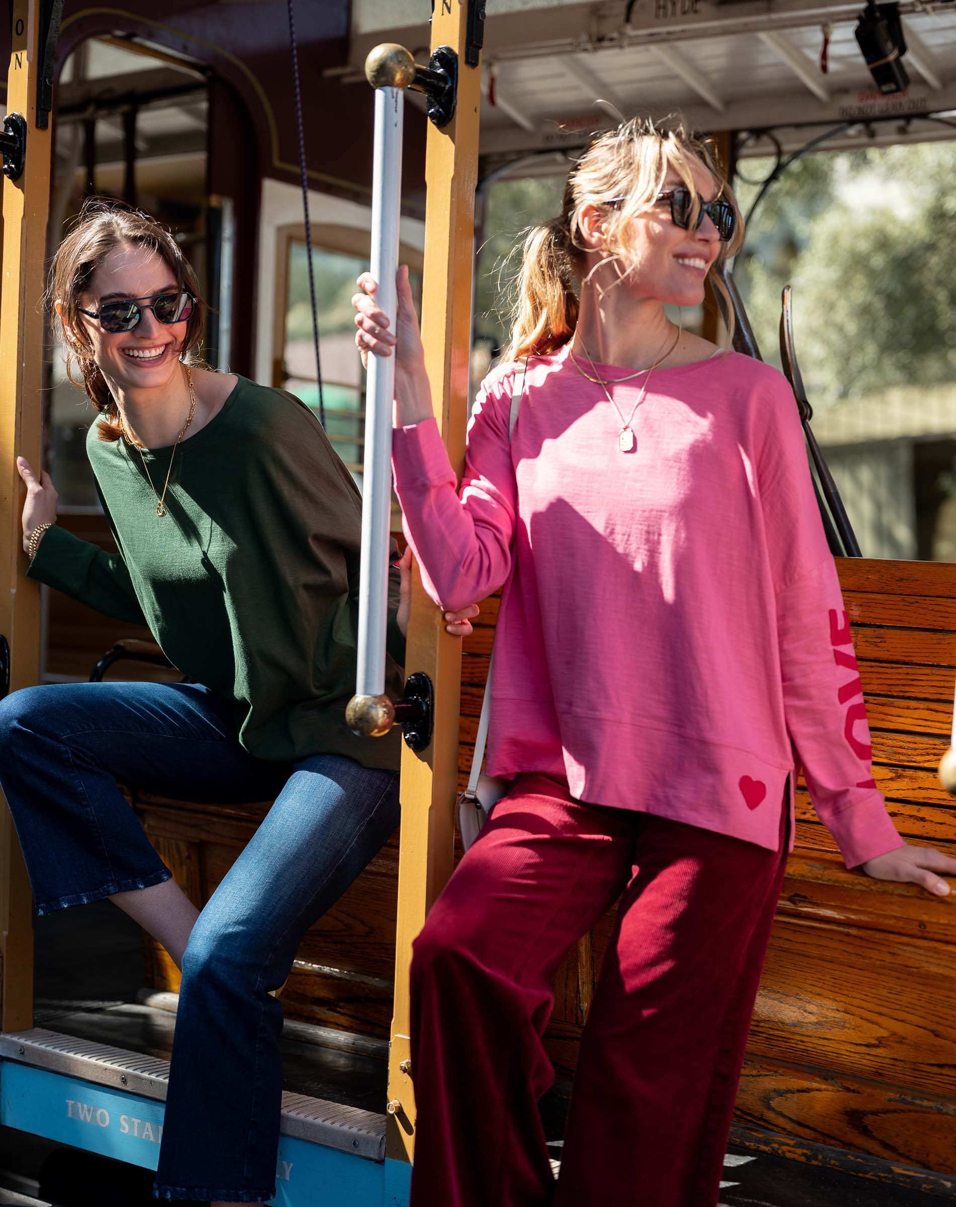 Two women on a trolley, one in a green top and blue jeans, the other in a pink sweatshirt and red pants.