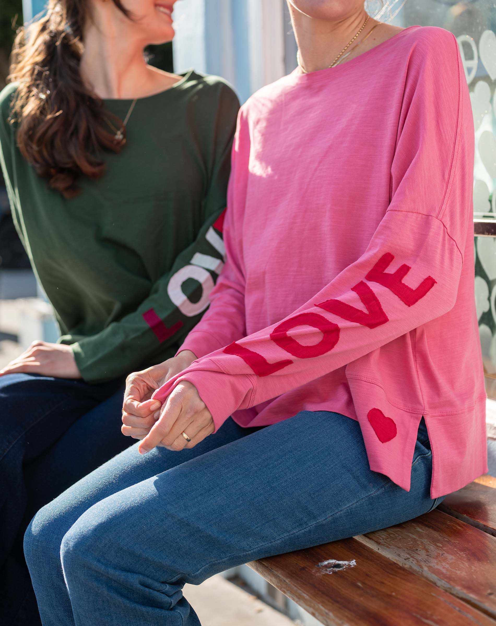 Two people sitting on a bench, one wearing a green shirt with 'LOVE' and the other in a pink sweater with 'LOVE' and heart designs.