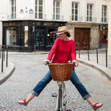 Woman in red sweater and blue jeans riding a bicycle with a basket on a city street.
