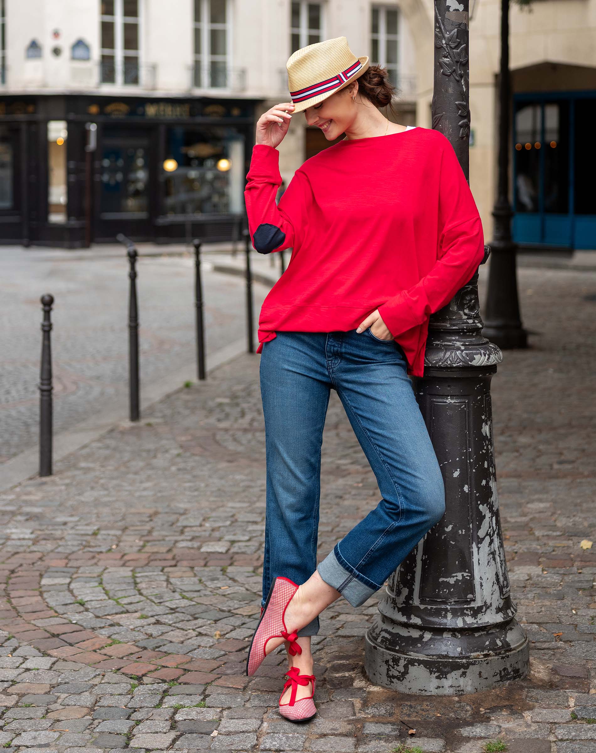 Woman in red sweater and blue jeans leaning against a lamp post on a street.