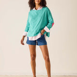 Woman wearing a teal sweater with white cuffs and hem, denim shorts, and brown sandals on a plain background