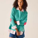 Woman wearing a teal sweater with pink elbow patches and denim shorts on a beige background