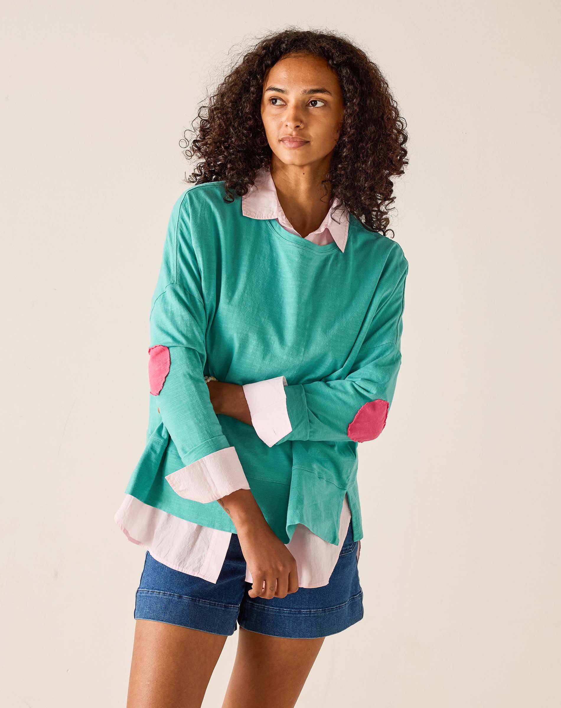 Woman wearing a teal sweater with pink elbow patches and denim shorts on a beige background